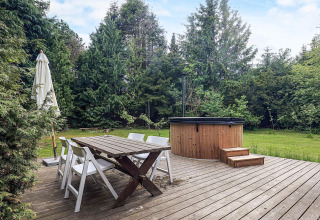 Wooden deck with dining table, chairs, parasol, and outdoor hot tub surrounded by trees in Idestrup.