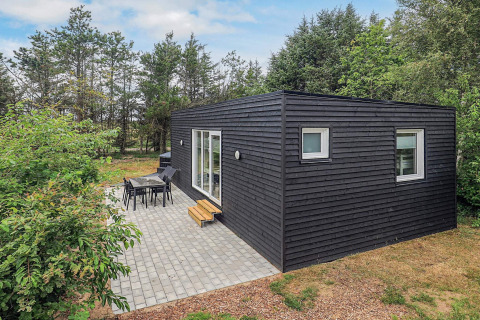 Cabaña negra moderna en Pandrup, Dinamarca, con terraza y muebles de jardín rodeada de árboles verdes.