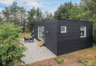 Cabaña negra moderna en Pandrup, Dinamarca, con terraza y muebles de jardín rodeada de árboles verdes.