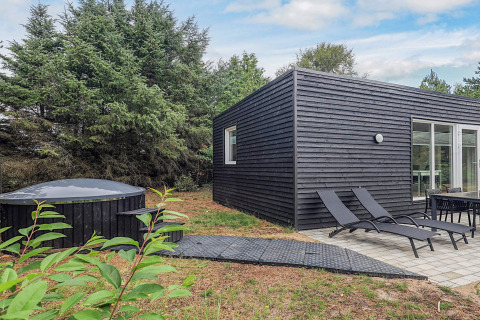 Vista exterior de una cabaña negra de vacaciones con tumbonas, mesa de comedor y jacuzzi cubierto en zona verde.
