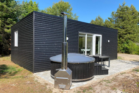 Cabaña negra de madera con jacuzzi exterior redondo y terraza, rodeada de árboles en el parque vacacional para 5 personas en Pandrup.