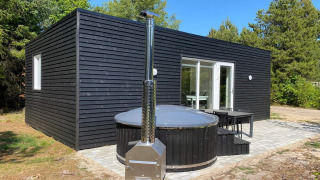 Black wooden cabin with round outdoor hot tub and patio, surrounded by trees at the 5-person holiday park home in Pandrup.