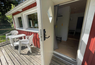 Open deur naar een rood houten huisje met terras, tuinstoelen en uitzicht op bos bij Furufjall Trail, Zweden.