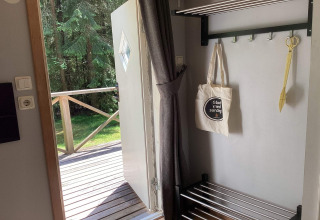 Open deur van een hut, uitzicht op houten terras en bos; canvas tas en gele schaar hangen binnen.