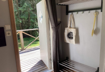 Puerta abierta en cabaña, vista a terraza de madera y bosque; bolsa de tela y tijeras colgadas.