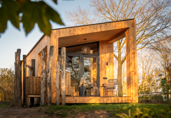 Wellness Duynzicht lodge met houten gevel, grote ramen, terras met stoelen en omringd door bomen in zonlicht.