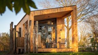Wellness Duynzicht lodge with wooden exterior, large windows, outdoor seating, and lush trees in sunlight.