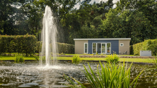 Knusse chalet met moderne uitstraling, grote fontein en vijver, omringd door groene bomen en struiken.