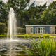Cosy cabin with a modern look, large fountain and pond in front garden, surrounded by lush green trees.