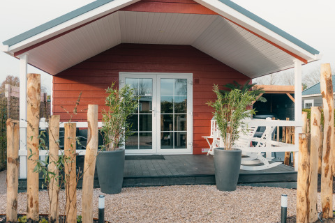 Cabaña roja con porche y plantas, Veluwe lounge + sauna en el parque vacacional De Boshoek, Países Bajos.