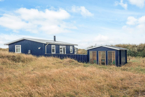 Lodge blu Seaside Bliss ad Albaek, circondato da erba alta e dune sotto un cielo parzialmente nuvoloso.
