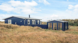 Blå træhytte kaldet Seaside Bliss i Ålbæk, omkranset af højt græs og klitter under en blå himmel.
