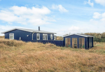 Lodge bleu Seaside Bliss à Albaek, entouré d’herbes hautes et de dunes sous un ciel partiellement nuageux.