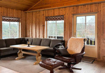 Salon de lodge avec murs en bois, canapé d’angle gris, fauteuil cuir et grandes fenêtres sur la nature.