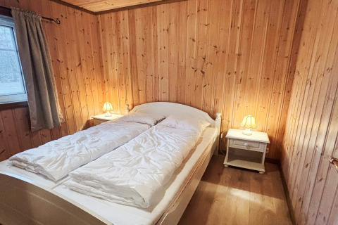 Soveværelse i lodge 76409 TROLLHYTTA i Danmark med dobbeltseng, trævægge og natborde med lamper.