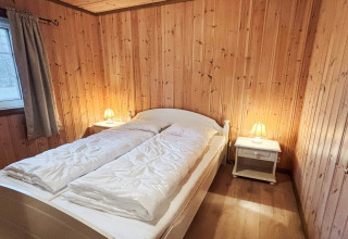 Soveværelse i lodge 76409 TROLLHYTTA i Danmark med dobbeltseng, trævægge og natborde med lamper.