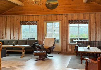 Salon chaleureux de la cabane 76409 TROLLHYTTA au Danemark, doté de meubles en bois et de grandes fenêtres.