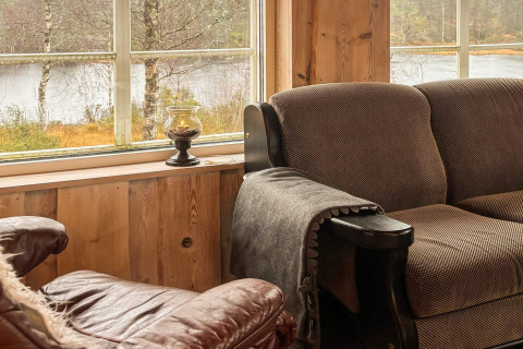 Hyggekrog med brun sofa og lænestol ved store vinduer med udsigt over søen i 76409 TROLLHYTTA-lodgen.