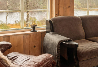 Hyggekrog med brun sofa og lænestol ved store vinduer med udsigt over søen i 76409 TROLLHYTTA-lodgen.