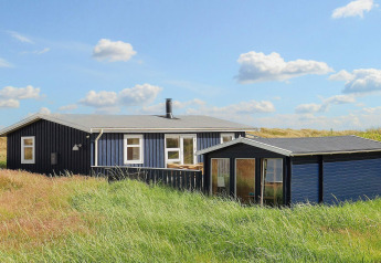Sort træhytte med store vinduer i klitlandskab under blå himmel nær havet i Albaek, Danmark.