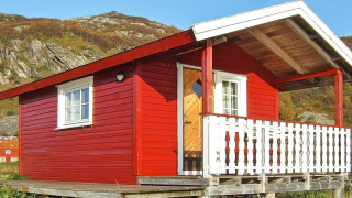 Red cabin named ENGVIK HYTTA 83699 with a white porch, set on wooden stilts in a scenic, hilly landscape.