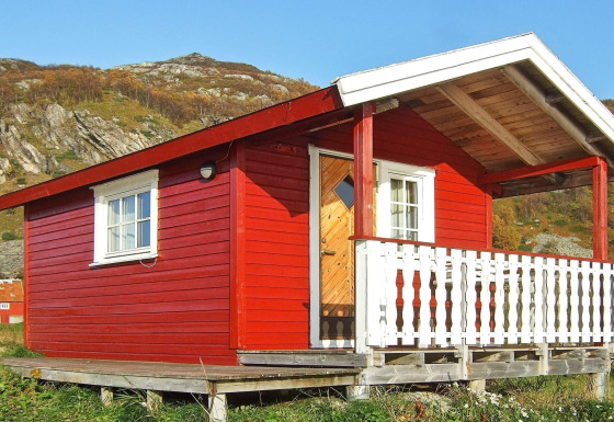 Casetta rossa chiamata ENGVIK HYTTA 83699 con veranda bianca, situata su palafitte in un paesaggio collinare.