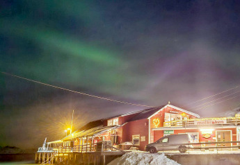 Nachtfoto van Engvik Hytta 83699 in Zweden met noorderlicht boven een besneeuwde en verlichte hut.