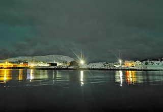 Vista notturna di baite e colline innevate in Svezia, illuminate da lampioni riflessi sull’acqua del lago.