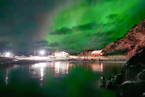 Immagine dell’aurora boreale sopra Engvik Hytta 83699 in Svezia, con cabine illuminate e riflessi sull’acqua.