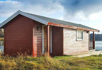 Cabaña roja de madera junto a un lago en Suecia, rodeada de césped y colinas bajo un cielo parcialmente nublado.