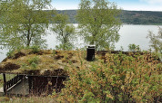 Chalet with a grassy roof by the lakeshore in Sweden, surrounded by trees and overlooking the water.