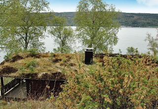 Chalet con tetto erboso vicino al lago in Svezia, circondato da alberi e con vista sull’acqua.