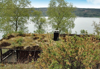 Chalet with a grassy roof by the lakeshore in Sweden, surrounded by trees and overlooking the water.