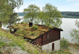 Traditionel svensk bjælkehytte ved søbred med græstag, træer i nærheden og udsigt til vand.