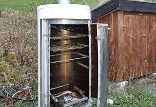 Fumoir extérieur avec porte ouverte et grilles métalliques, à côté d’un abri en bois à Surna.