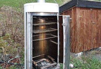 Fumoir extérieur avec porte ouverte et grilles métalliques, à côté d’un abri en bois à Surna.