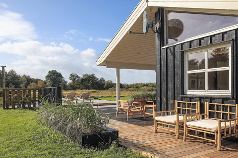 Terraza de madera con asientos al aire libre junto a una cabaña negra, rodeada de campo y naturaleza.