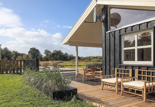 Terraza de madera con asientos al aire libre junto a una cabaña negra, rodeada de campo y naturaleza.