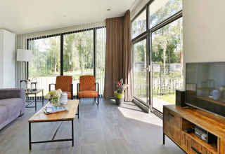 Moderne lodge woonkamer met grote ramen en zicht op de tuin in De Lochemse Berg vakantiepark.
