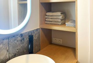 Modern bathroom with wooden shelves, folded towels, LED mirror, and power outlets at Lysdann lodge, Belgium.