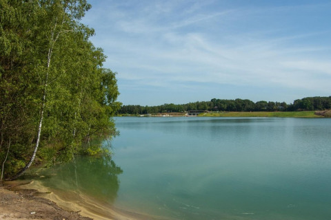Scenic view of the lake at Lysdann lodge in Belgium, with lush green trees and calm water under blue skies.