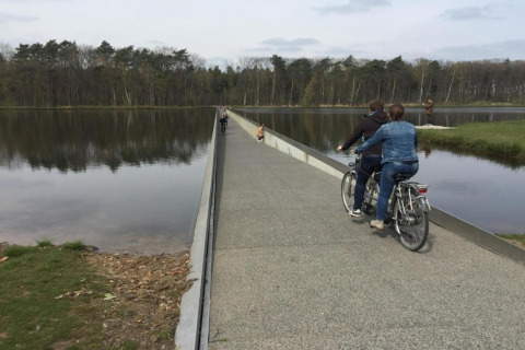Two people cycle on a submerged bridge crossing a lake at the Lysdann lodge in Belgium, with forest views.