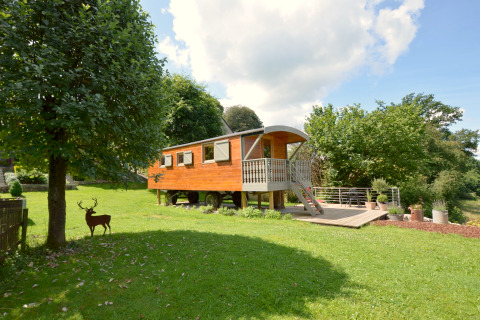 Casa móvil Manureva en césped verde con árboles, terraza de madera, plantas en macetas y naturaleza.