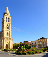 Iglesia con torre alta junto a una rotonda en un día soleado, rodeada de jardines y edificios clásicos.