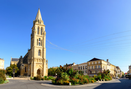 Kerk met hoge toren aan een rotonde op een zonnige dag, omgeven door bloemperken en klassieke gebouwen.