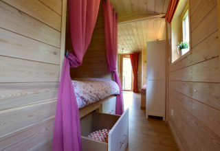 Vista interior de la casa móvil Manureva en Bélgica, con cama, cortinas rosas y paredes de madera.