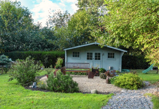 Jardín verde con arbustos, banco y casa móvil azul 'Manureva' situada en Bélgica bajo luz del sol.