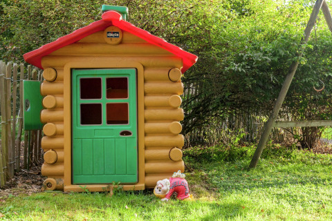 Casita de juegos estilo madera con puerta verde y techo rojo, letrero de bienvenida, en un jardín.