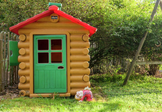 Casita de juegos estilo madera con puerta verde y techo rojo, letrero de bienvenida, en un jardín.