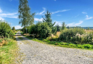 Camino de grava junto a la casa móvil Manureva en Bélgica, rodeado de arbustos bajo cielo azul claro.